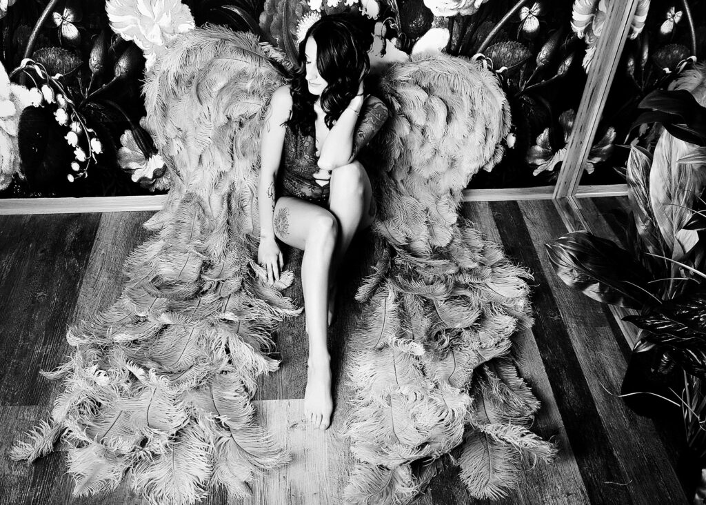 A woman in lace lingerie sits on a studio floor while wearing large feather angel wings after doing brazilian waxing milwaukee